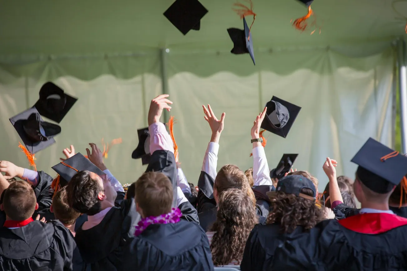A group of graduates in caps and gowns joyfully toss their hats into the air inside a tent. 