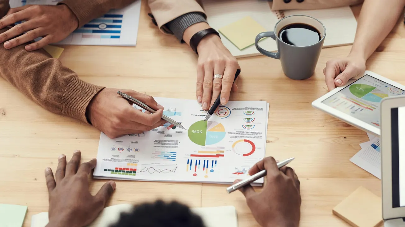 Hands of diverse individuals are gathered around a table with papers showing charts and graphs. A tablet and coffee cup are also visible, conveying teamwork.