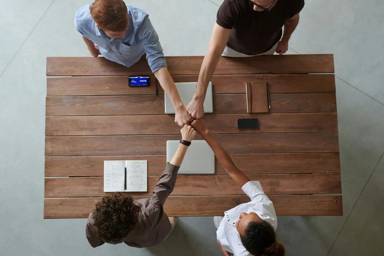 Four diverse individuals stand around a wooden table, fist-bumping in the center. Laptops, notebooks, and a phone suggest a collaborative workspace.