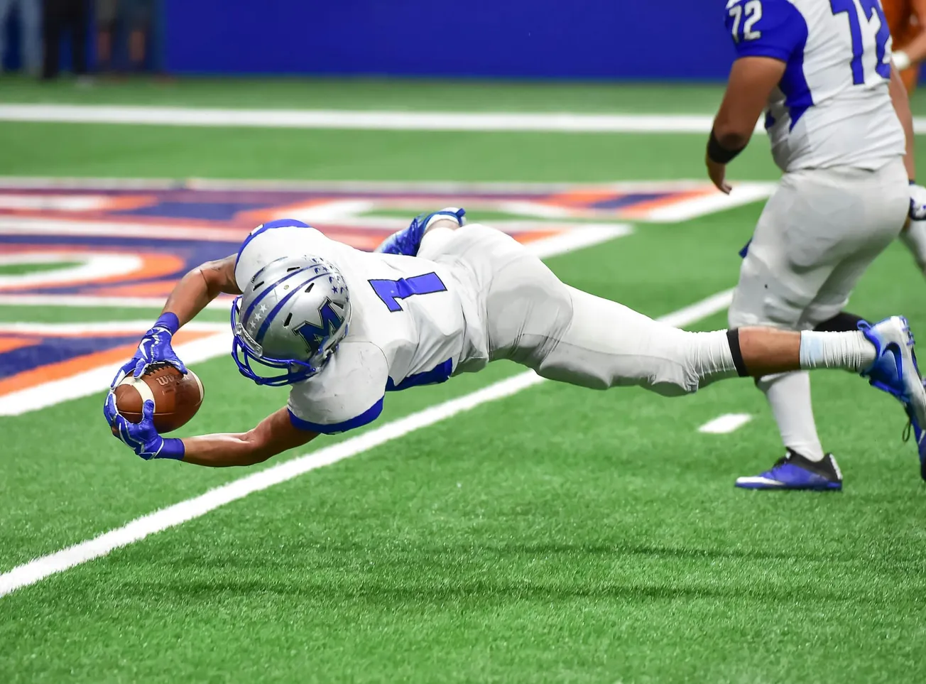 A football player in a white and blue uniform dives to catch a ball near the end zone line on a green field.