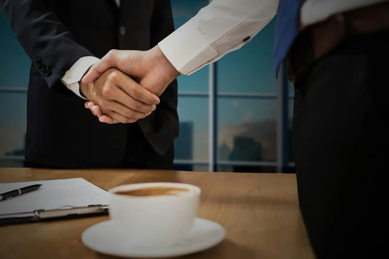 Two businesspeople in suits shake hands over a meeting table with a coffee cup and clipboard.