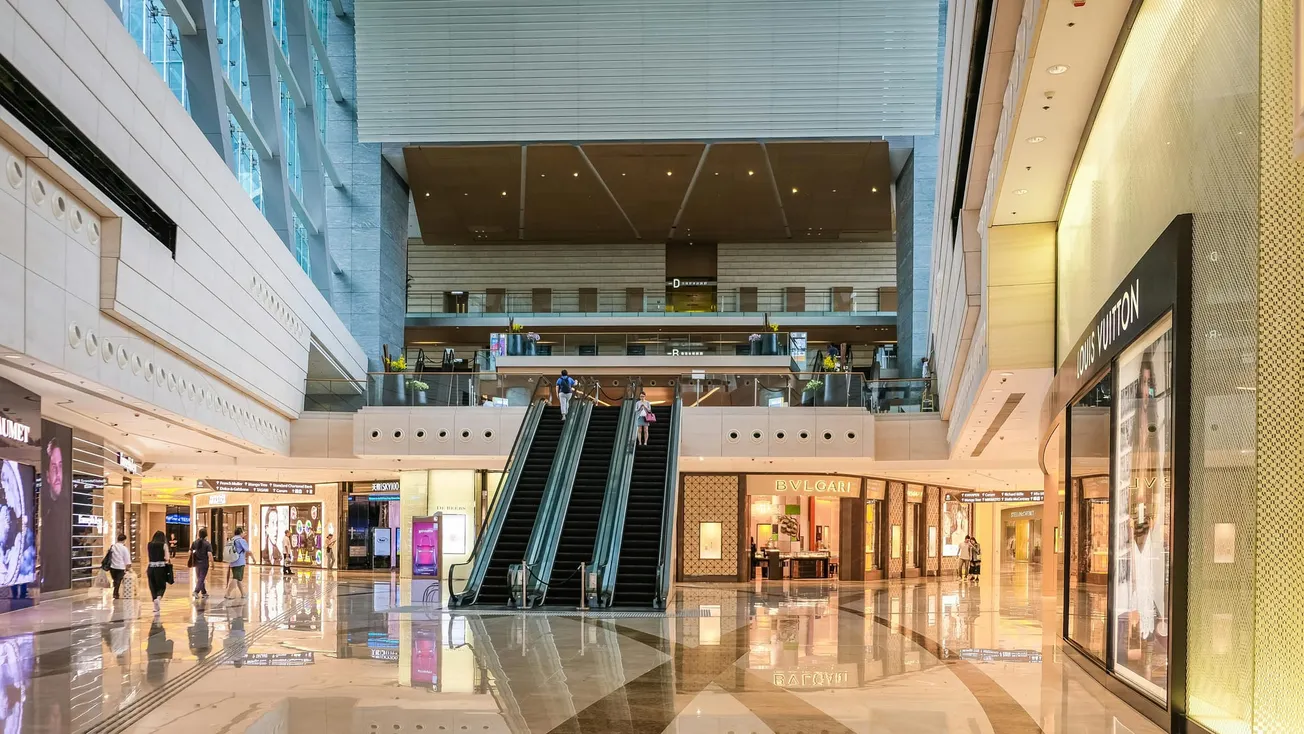 Spacious shopping mall interior with bright lighting and polished floors. Central escalator leads to upper level, surrounded by luxury brand stores.