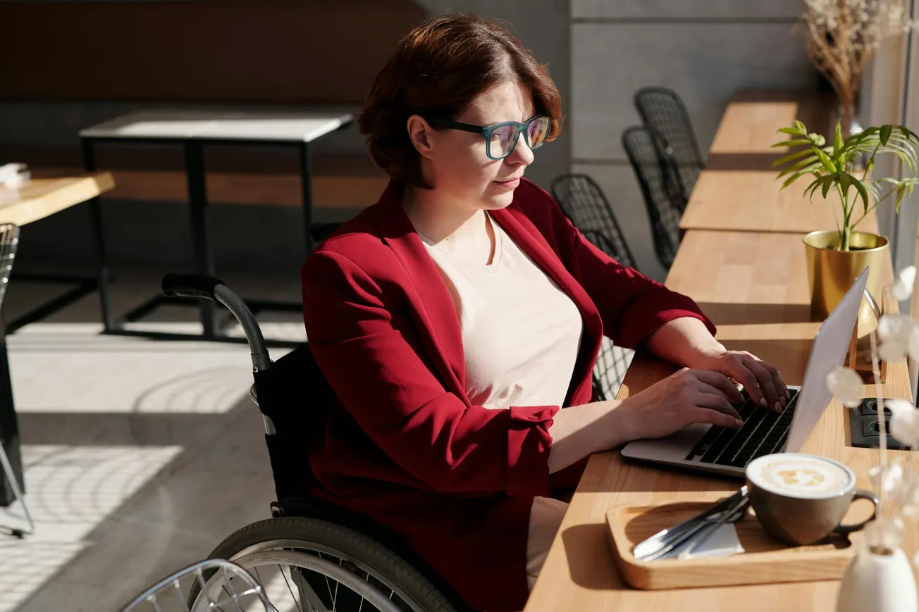 A woman in a red blazer and glasses works on a laptop at a wooden counter in a bright cafe. She sits in a wheelchair, focused, with a latte nearby.