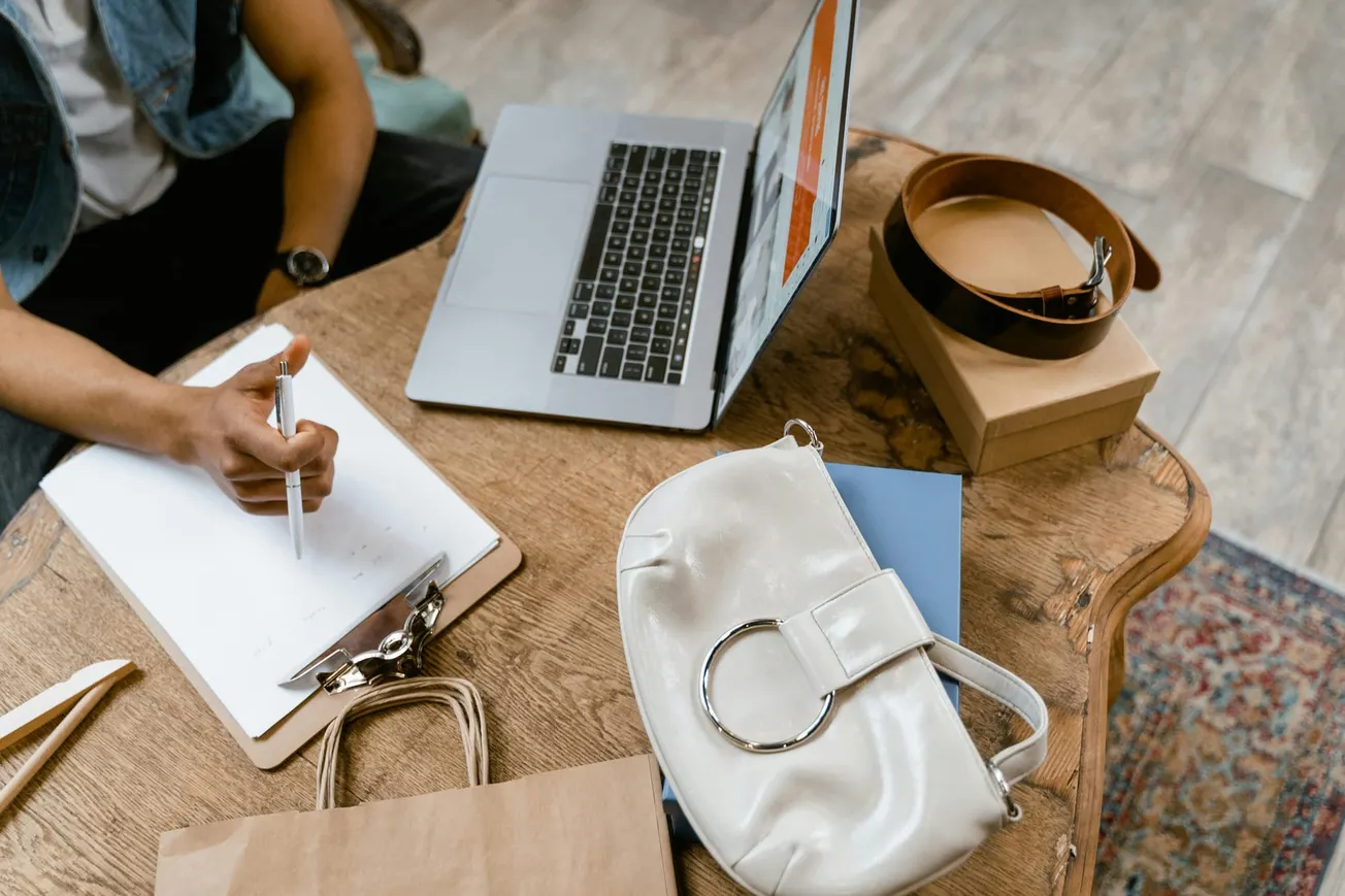 Person writing on a clipboard at a wooden table with a laptop, white handbag, and stacked gift boxes.