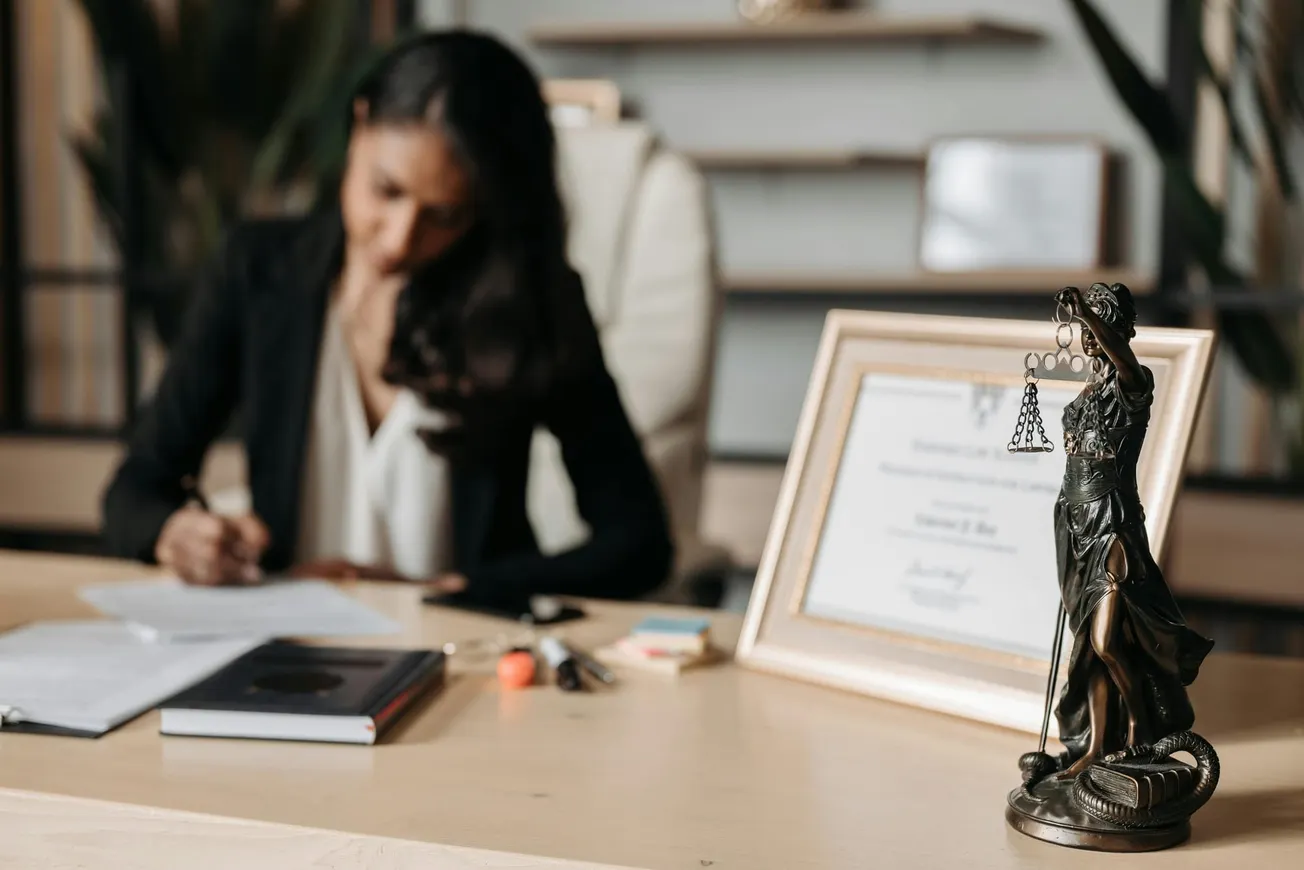 A woman in a black blazer writes at a desk with focus on a Lady Justice statue and framed certificate.