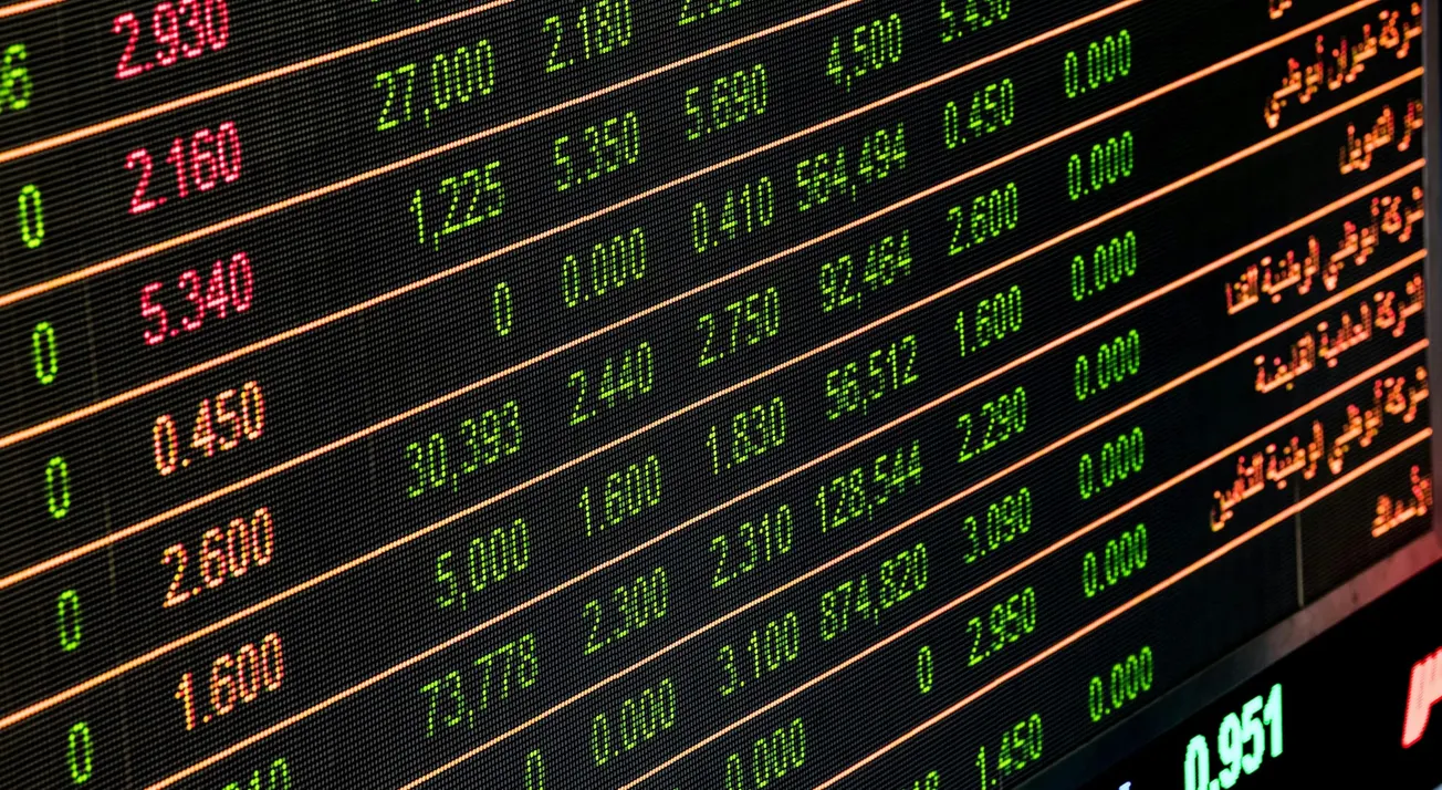 Close-up of a digital stock market board displaying numbers in green, red, and orange against a black background.