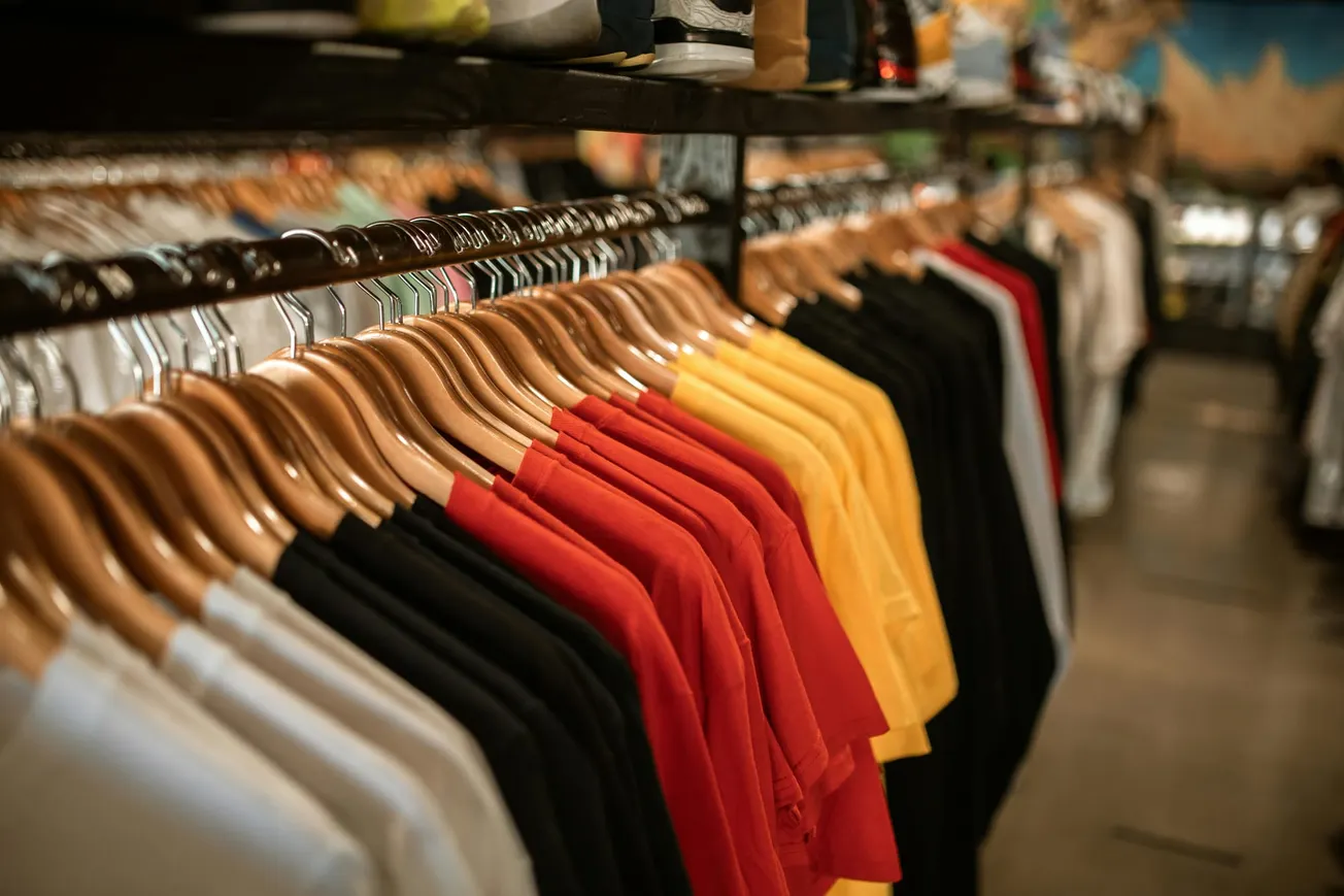 A row of colorful t-shirts in white, black, red, and yellow hang neatly on wooden hangers in a clothing store.