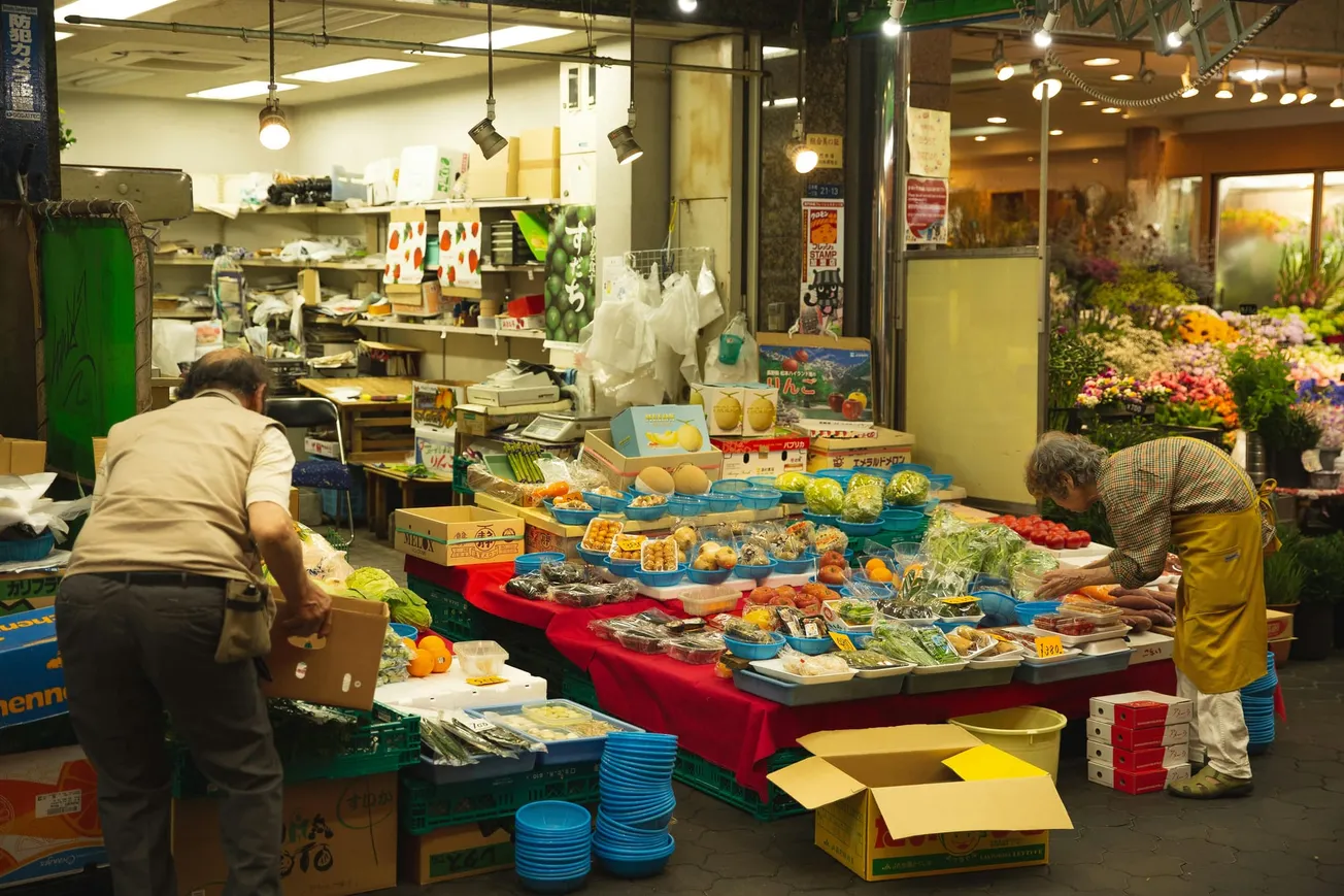 A vibrant market scene showing two people organizing fresh produce on tables. The stands are laden with fruits, vegetables, and melons. 