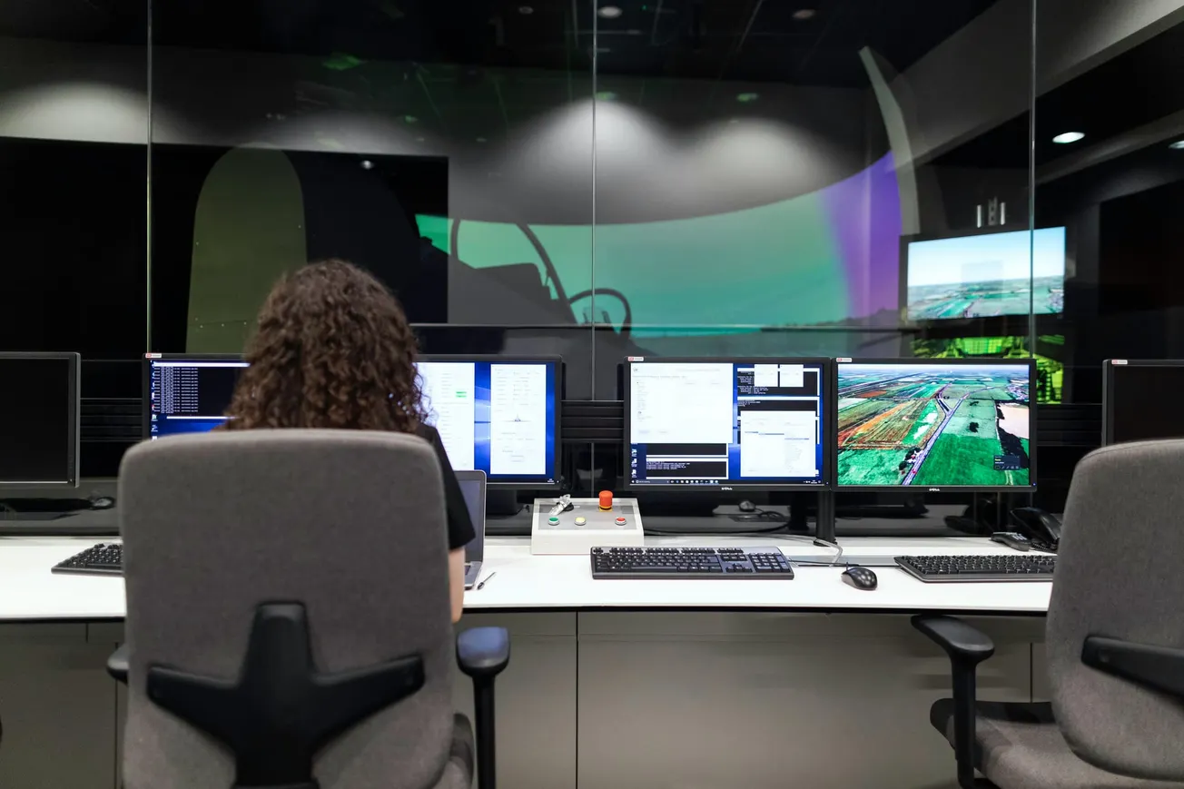 A person with curly hair sits at a control desk in front of multiple monitors displaying software and aerial images, in a control room.