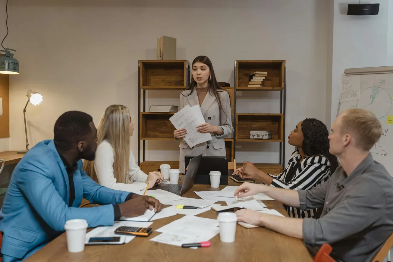 A woman in a blazer stands, holding documents, leading a meeting with four colleagues seated at a table covered with papers and coffee cups.