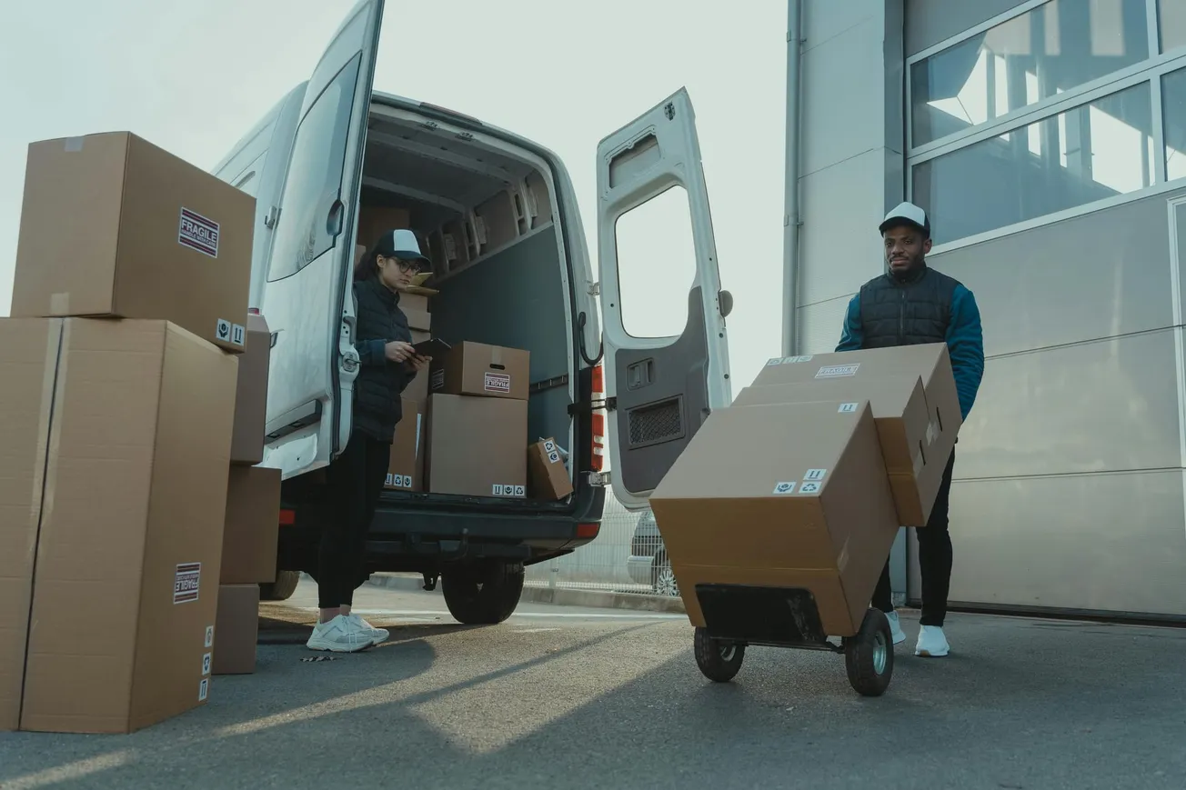 Two people load cardboard boxes into a van. One person uses a dolly, while the other checks items.