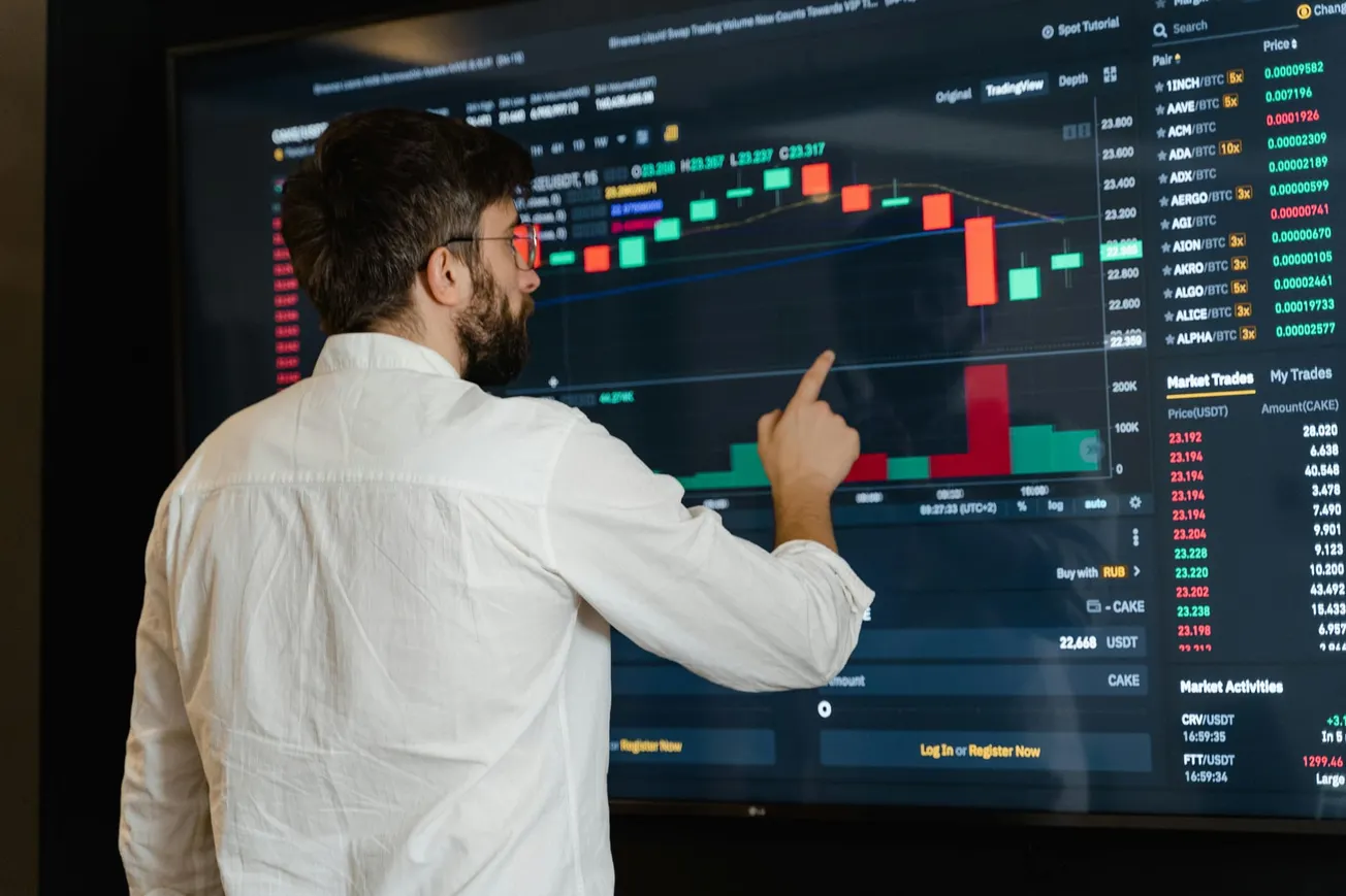 A man in a white shirt analyzes a digital trading chart on a large screen, pointing at a candlestick graph displaying cryptocurrency market data.