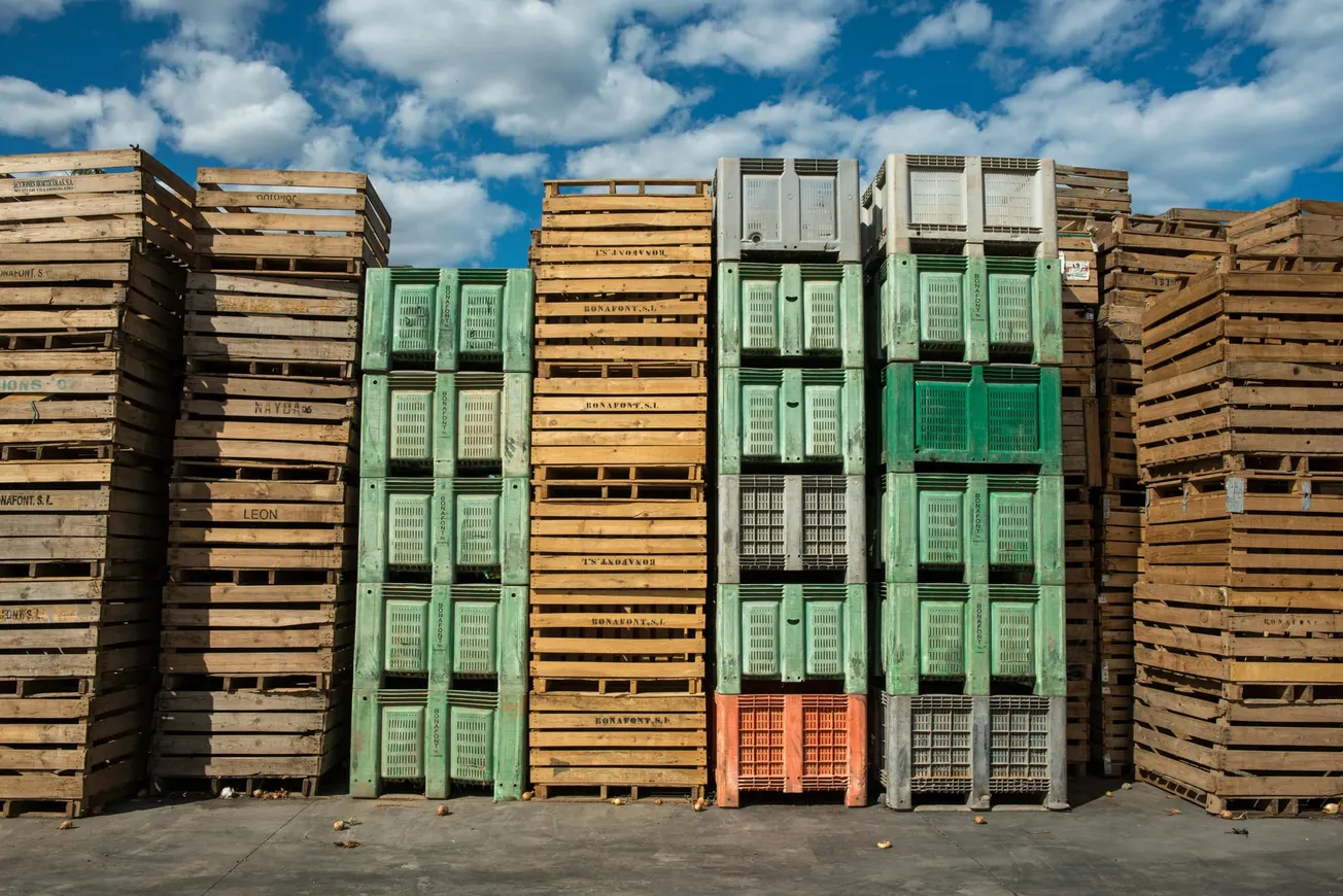 Stacks of wooden and plastic crates arranged outdoors under a blue sky with clouds. 