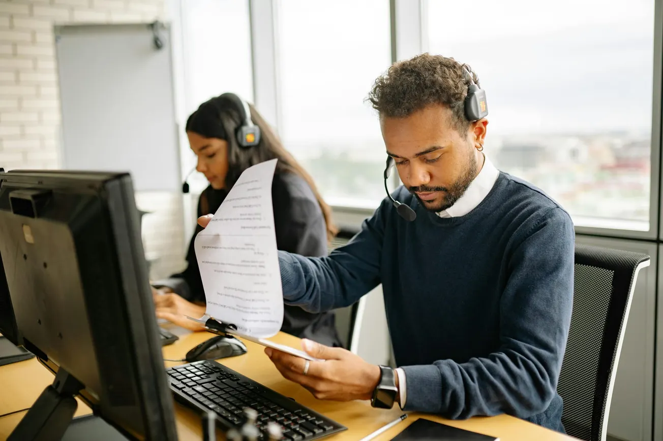 Man in an office wearing a headset reads a document at a computer. A woman in a headset works at the next desk.