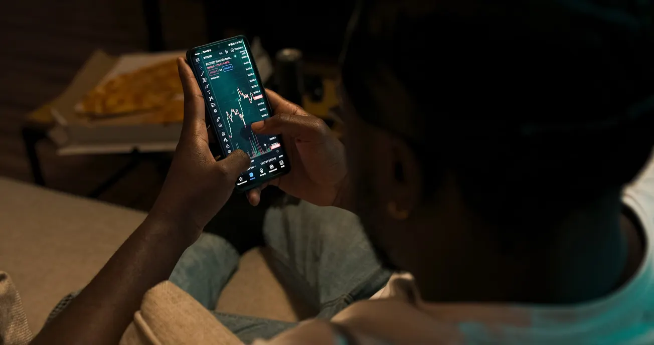 A person holds a smartphone displaying a stock market chart, suggesting financial trading. Nearby, a pizza box and drink convey a casual atmosphere.