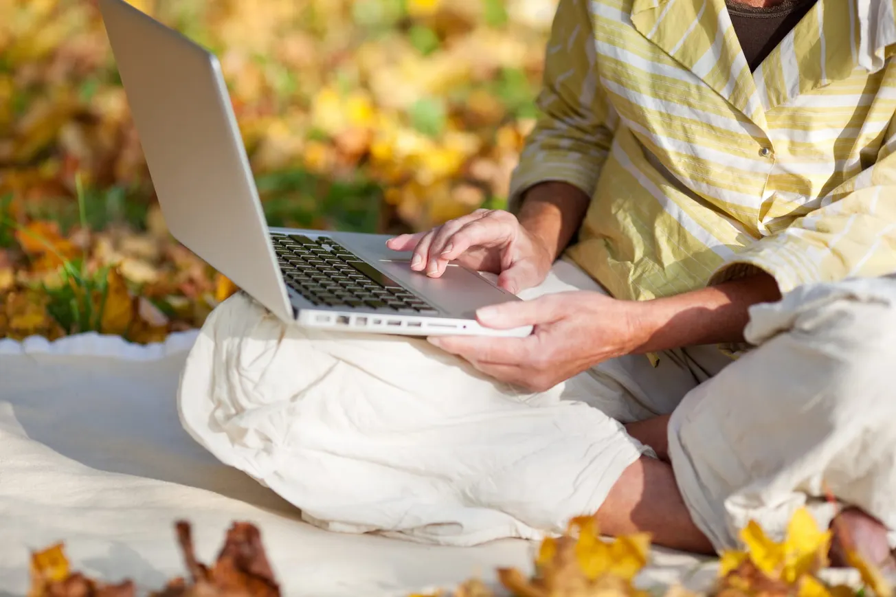 Person in a yellow striped shirt and white pants sits cross-legged on a blanket, using a laptop outdoors. Surrounded by autumn leaves, the scene is calm and focused.