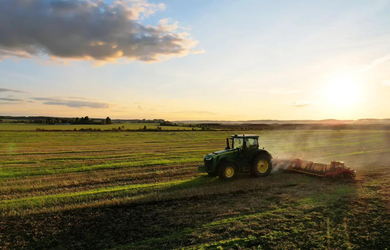 A green tractor plows a vast, sunlit field under a blue sky with scattered clouds. The sun sets on the horizon, casting a warm glow across the landscape.