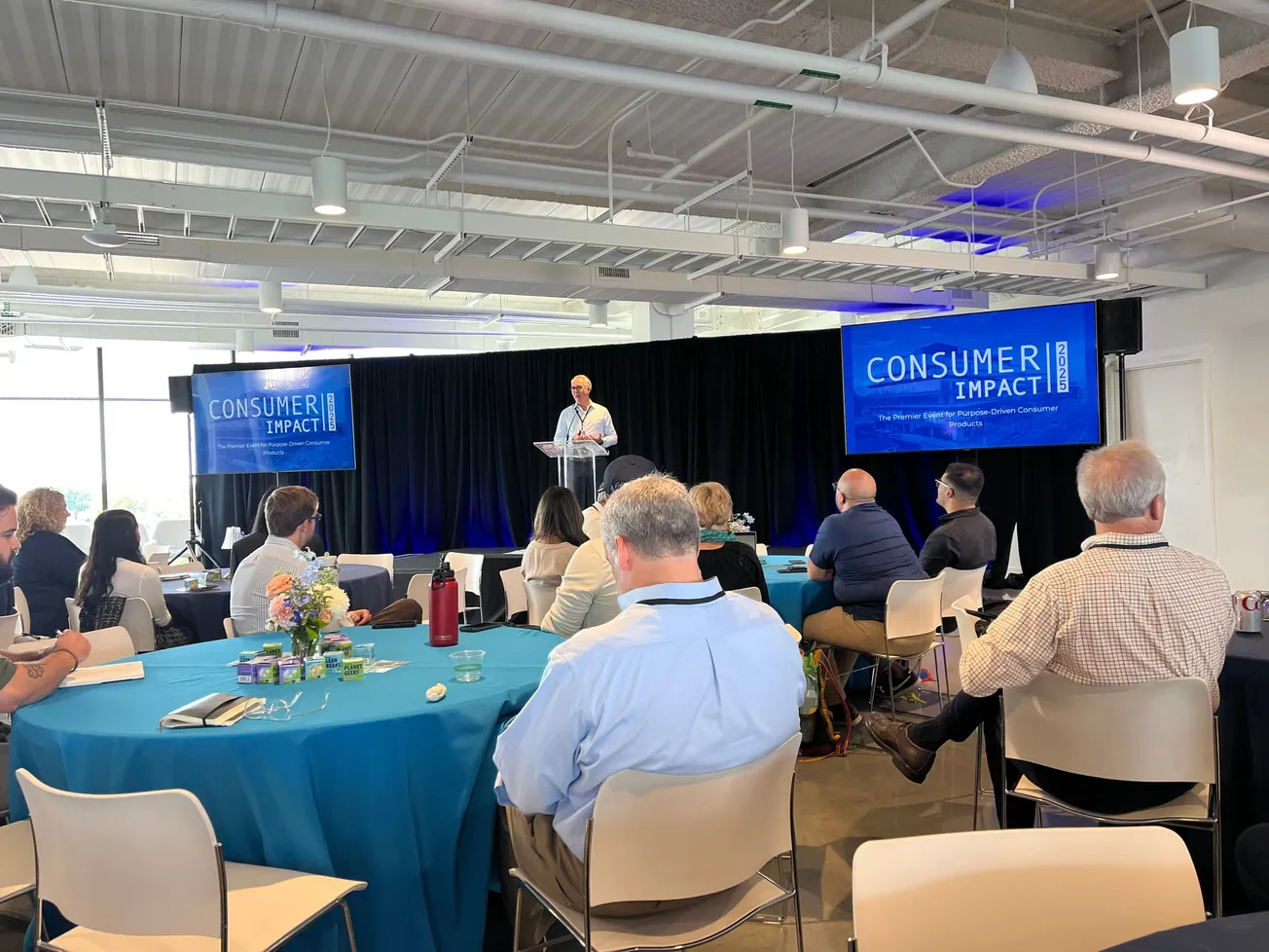 A speaker stands at a podium on stage in a conference room with seated attendees. Two screens display "Consumer Impact." 