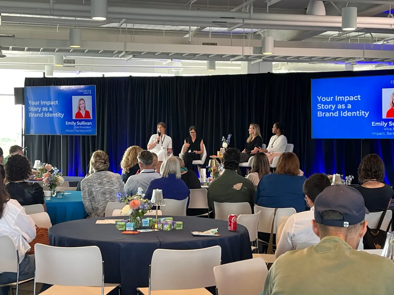 Panel discussion with four women seated on stage before an audience. Blue screens display "Your Impact Story as a Brand Identity."