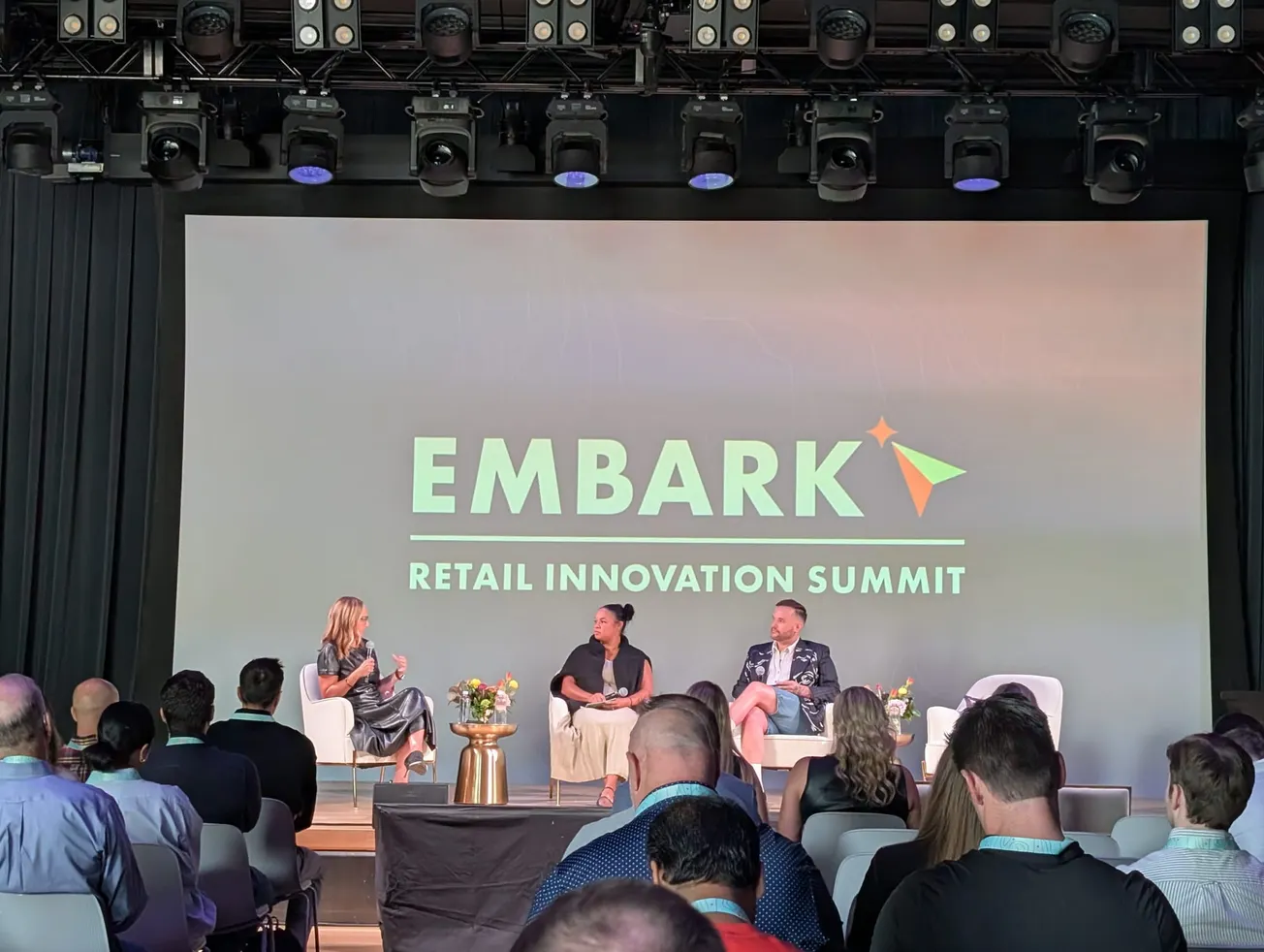 A panel of three people is seated on stage at a retail innovation summit, with "Embark" displayed behind them. Audience members are visible, listening attentively.