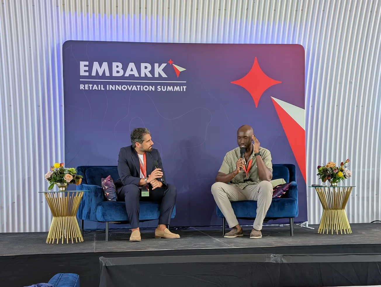 Two men are seated on stage at the Embark Retail Innovation Summit. They are engaged in a discussion, holding microphones.