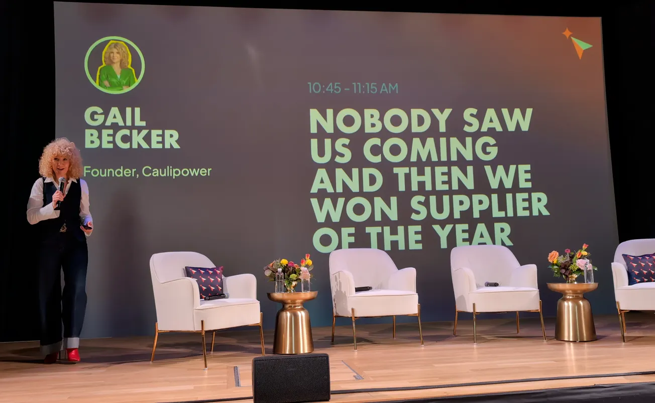 A woman speaks on stage in front of a large screen displaying the text, "Nobody saw us coming and then we won Supplier of the Year."