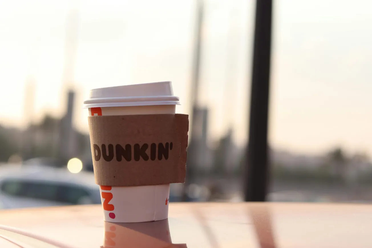 A Dunkin' coffee cup with a cardboard sleeve sits on a car roof with a soft-focus urban background, capturing a calm morning vibe.
