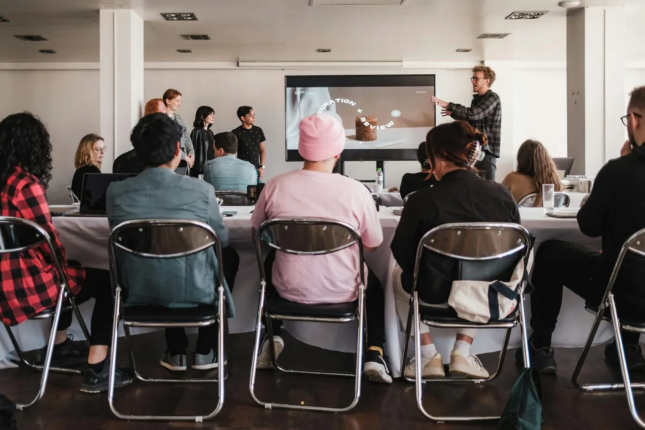 A group of people in a classroom setting listens to a presentation. A speaker gestures towards a screen. Attendees sit at a table, creating an engaged atmosphere.