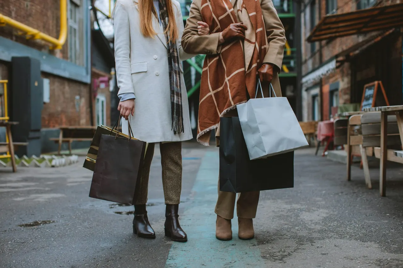 Two people in coats and scarves walk arm-in-arm on a city street, carrying shopping bags. The scene conveys a cozy, autumn shopping vibe.