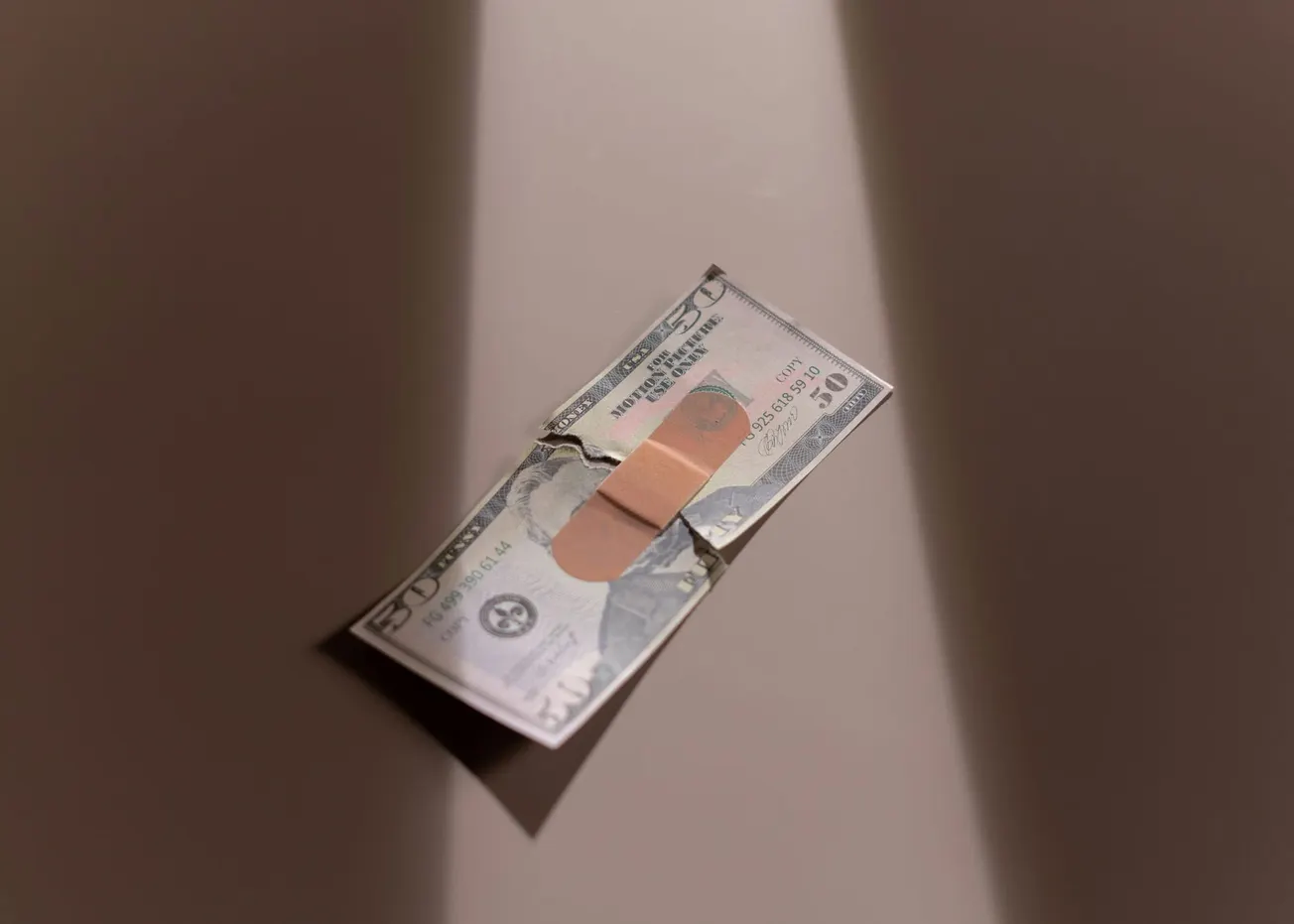 A torn $50 bill is mended with a band-aid, lying on a flat surface. Soft lighting creates a dramatic shadow.