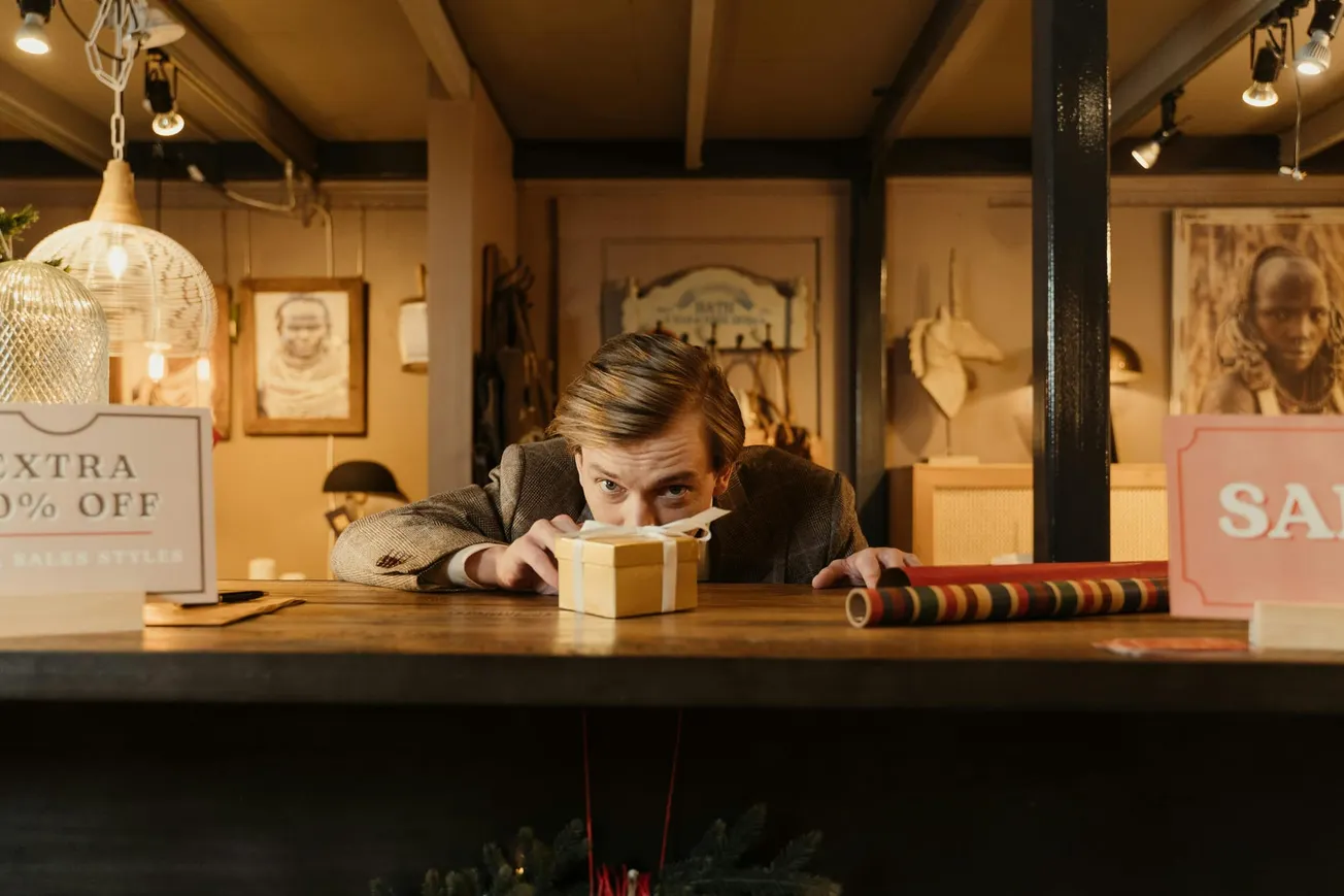 A man in a vintage shop peers over a counter at a small wrapped gift. Sale signs and hanging lights create a cozy, curious atmosphere.