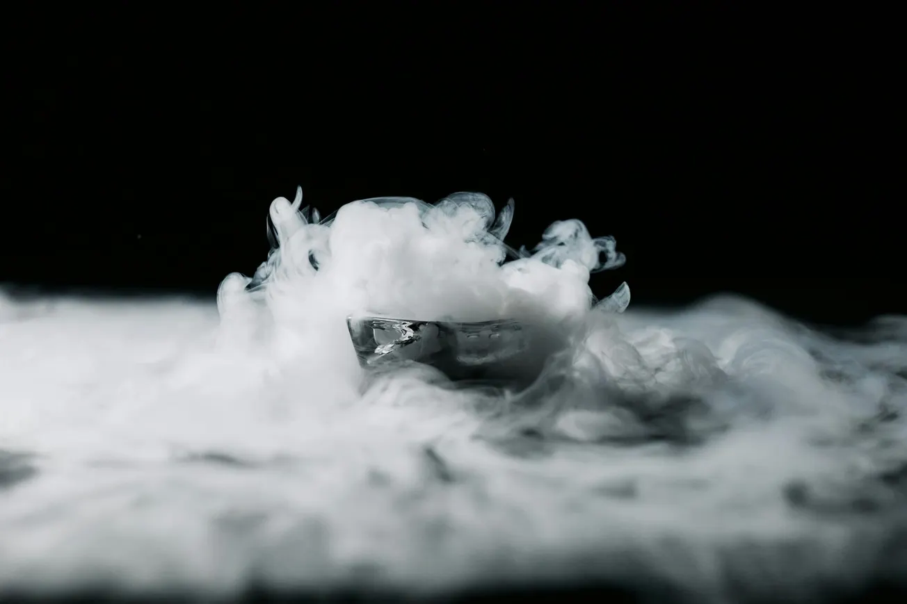 A glass sits amidst swirling white smoke on a dark background, creating a mysterious and dramatic atmosphere.