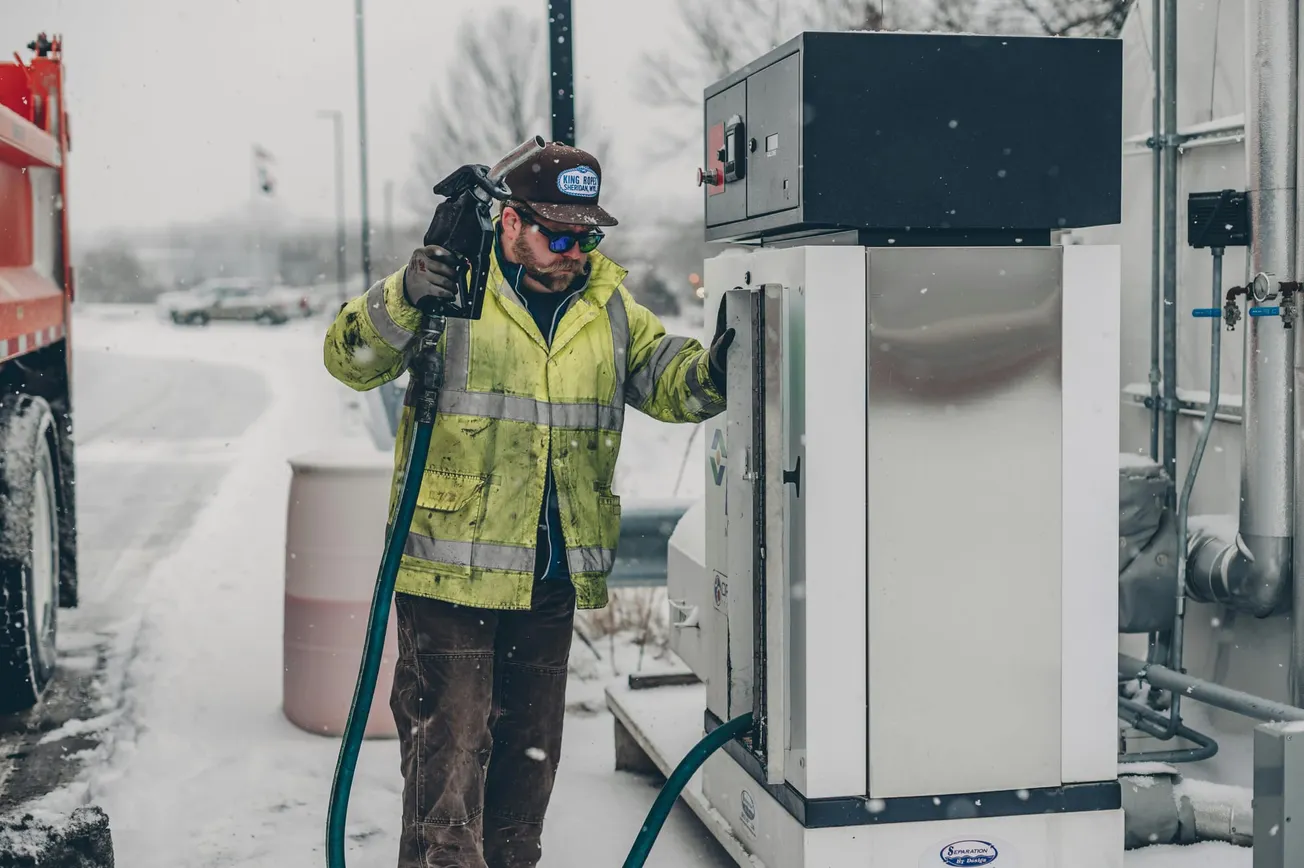 A worker in a bright yellow jacket and cap handles fuel equipment at a snowy gas station, conveying a sense of cold, diligence, and focus.