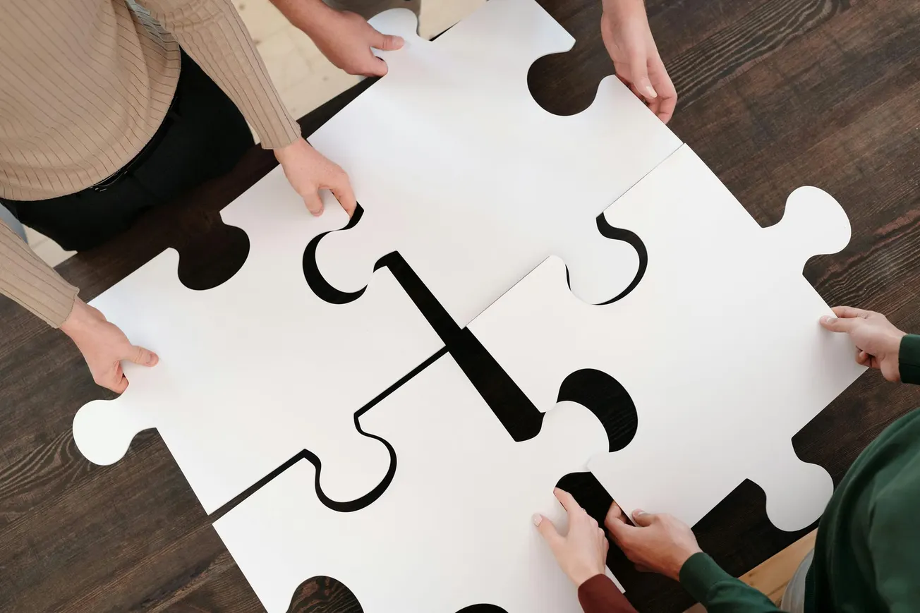 Four people collaboratively assembling large white puzzle pieces on a dark wooden table.