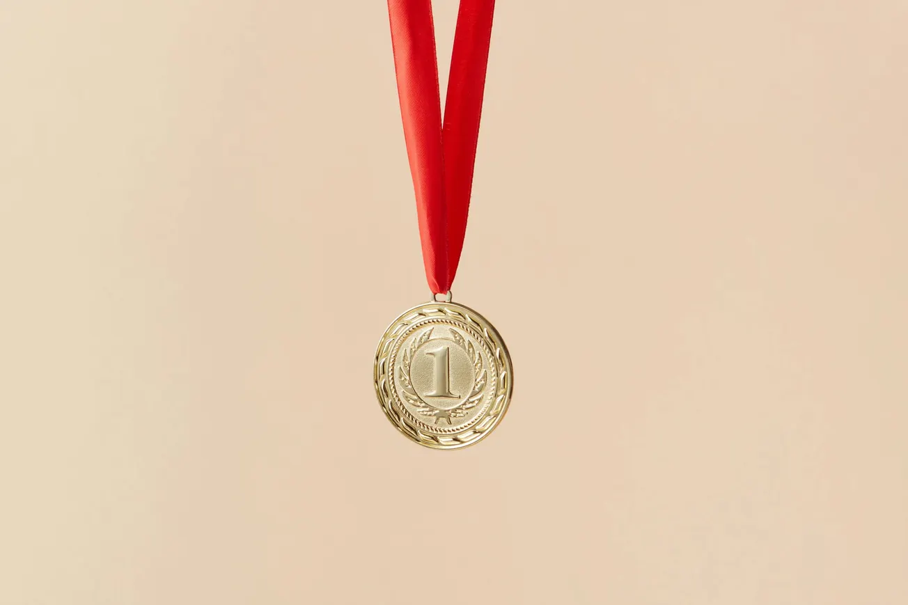 Gold medal with a number "1" embossed, hanging from a red ribbon on a light beige background, conveying achievement and victory.