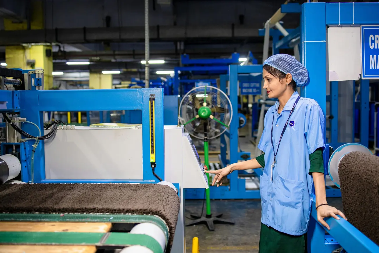 A factory worker in a blue uniform monitors machinery on a production line. The setting is a brightly lit manufacturing facility.