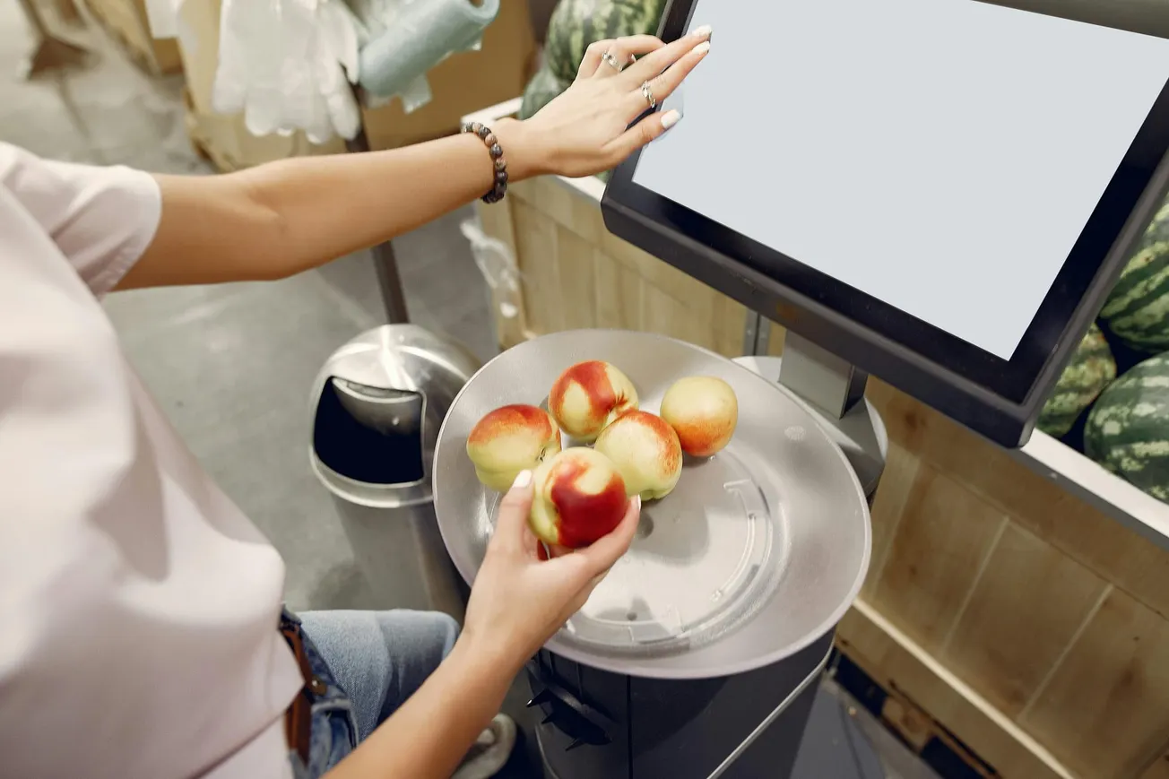 A person at a grocery store self-checkout scans red and yellow apples on a scale. Nearby, a touchscreen displays options. The scene is organized and casual.