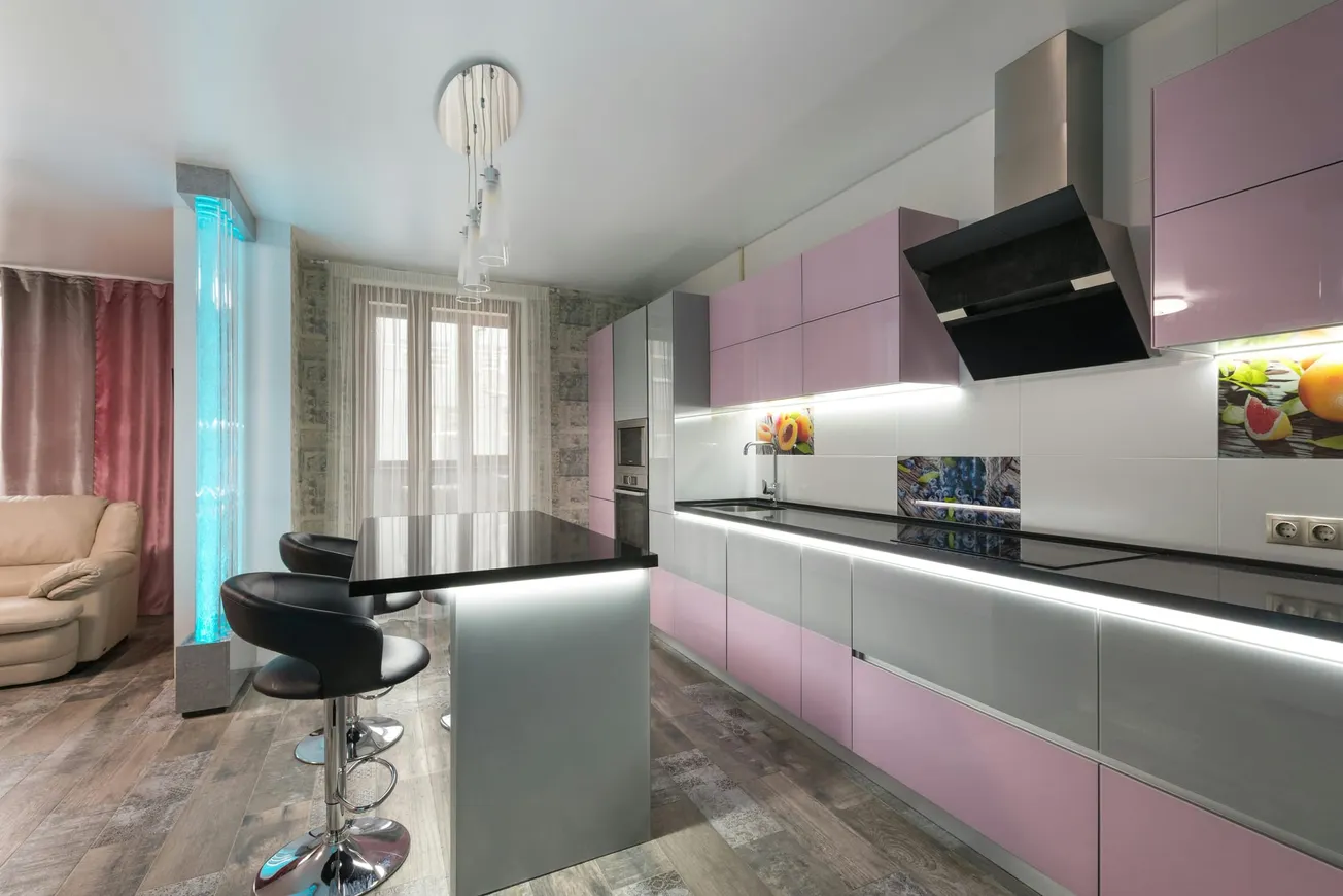 Modern kitchen with pastel pink and gray cabinets, illuminated by under-cabinet lighting. Features a sleek black countertop and three bar stools.