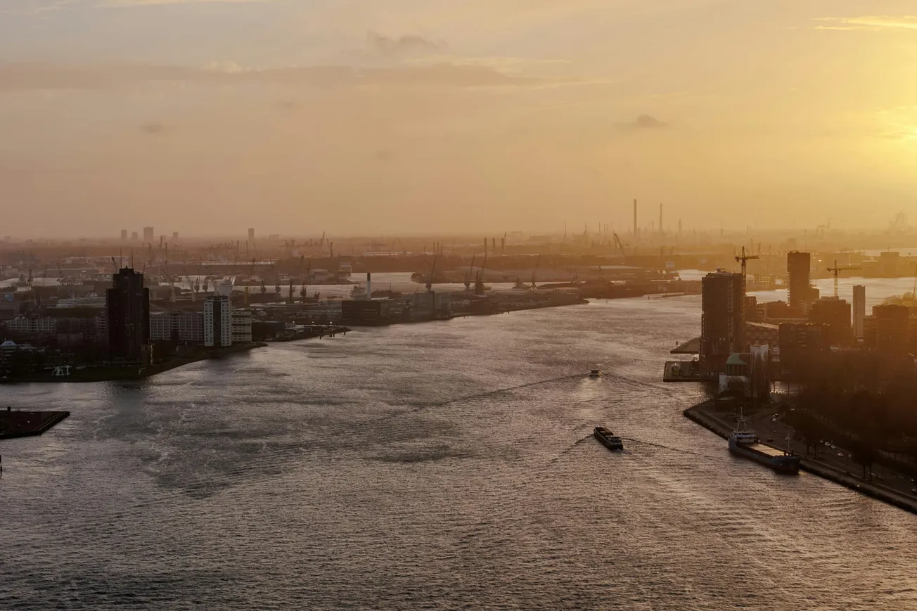 A serene river scene at sunset with scattered boats and industrial buildings on both sides. The sky is warm and hazy.
