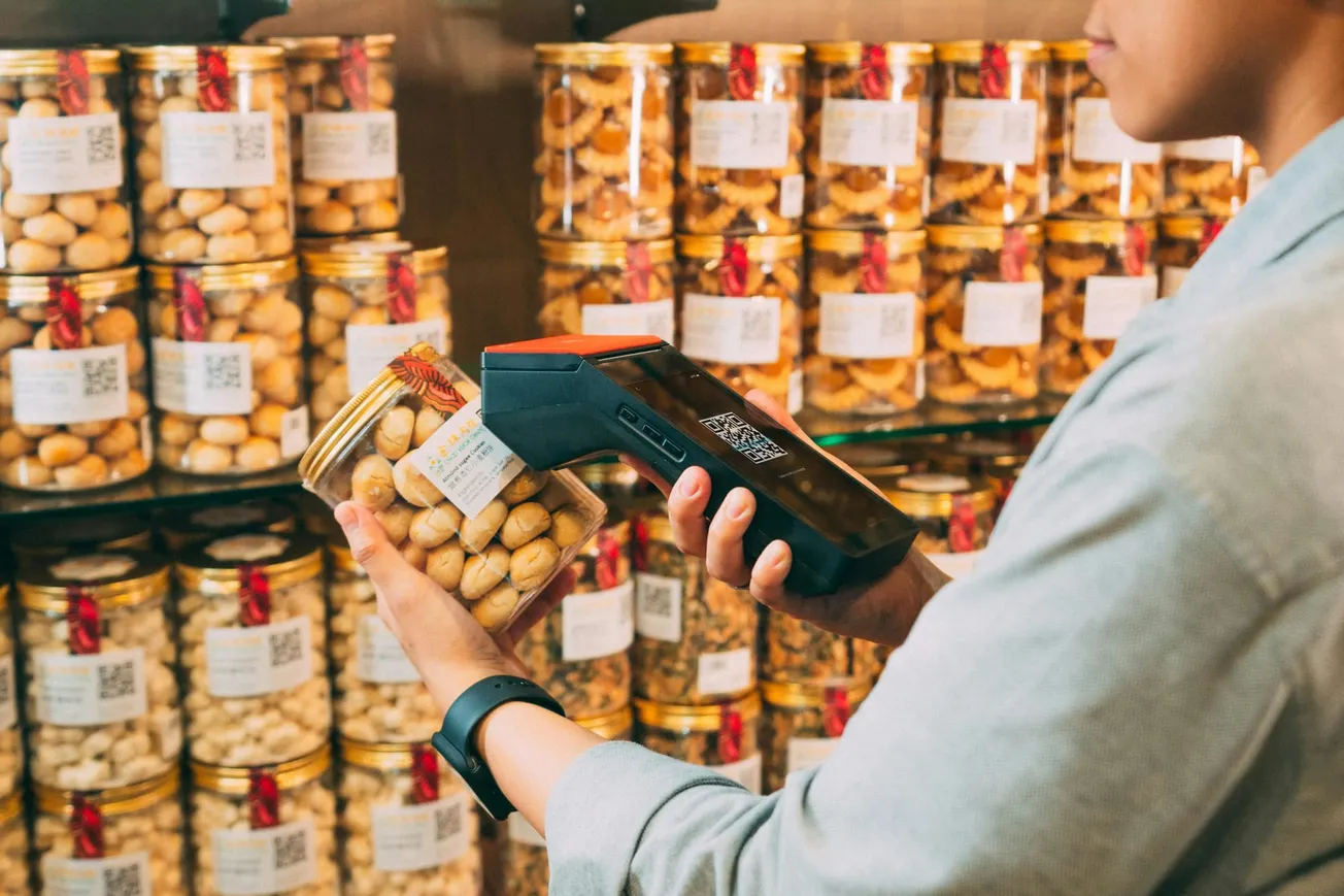 A person in a store scans a jar of snacks with a handheld device. Shelves filled with similar jars are in the background, creating a busy retail setting.
