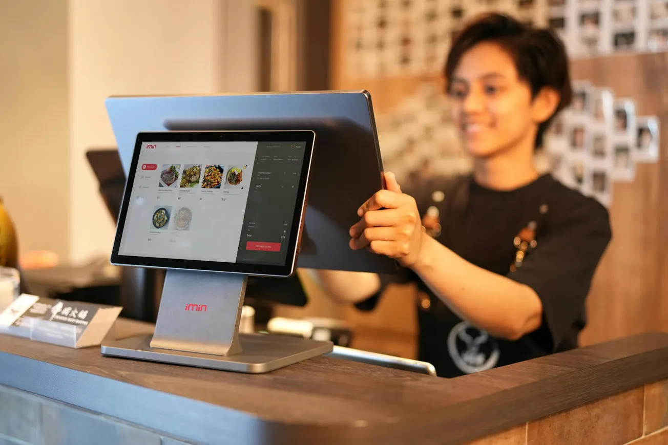 A smiling person stands behind a counter using a touchscreen point-of-sale system displaying menu items.