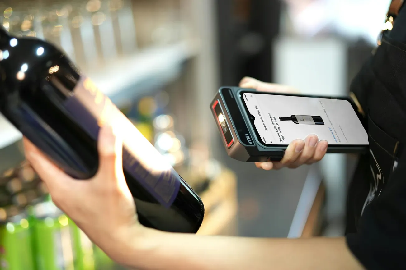 A person scans a wine bottle with a handheld device in a store, displaying product details on the screen.