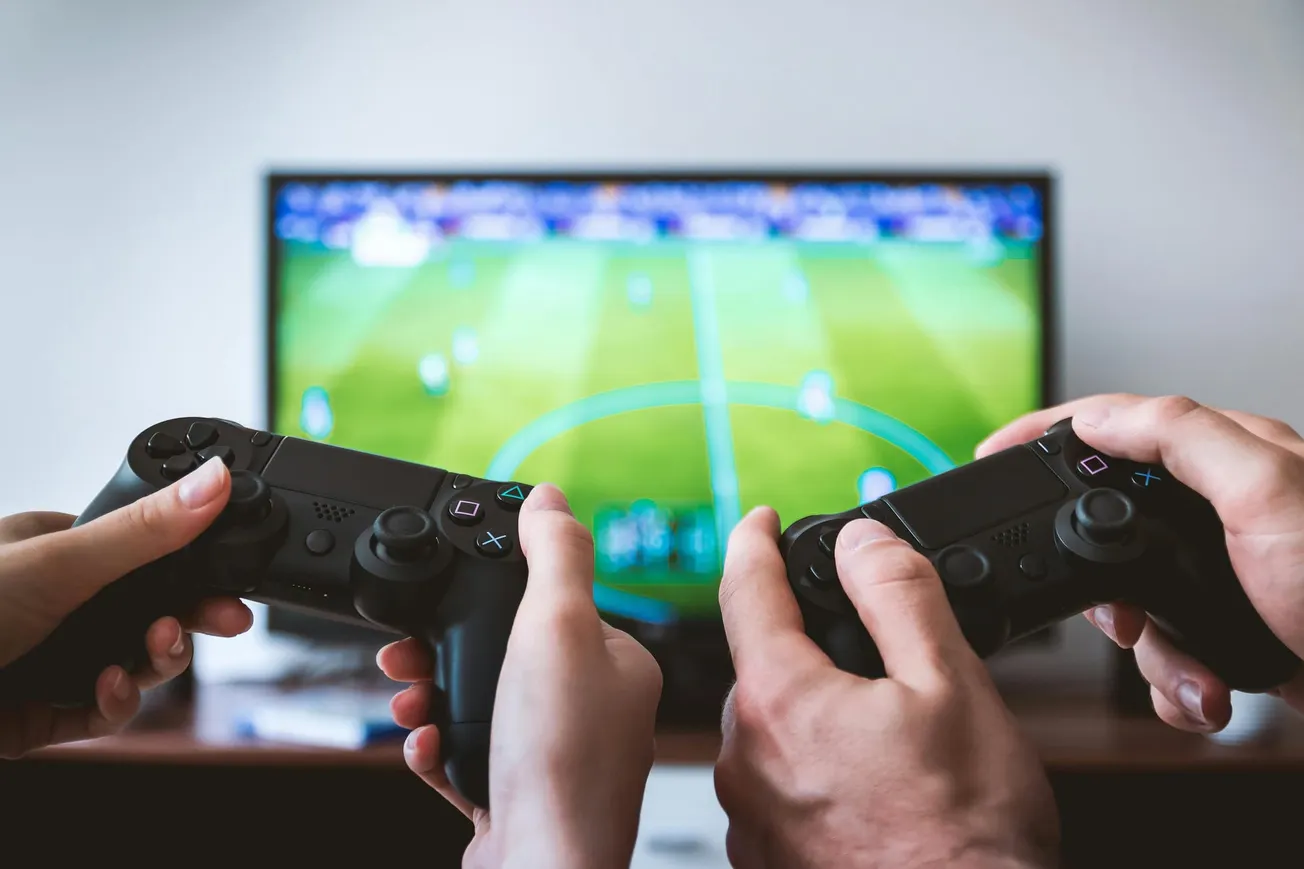 Two hands holding game controllers in front of a TV screen displaying a soccer game, suggesting excitement and focus in a gaming session.