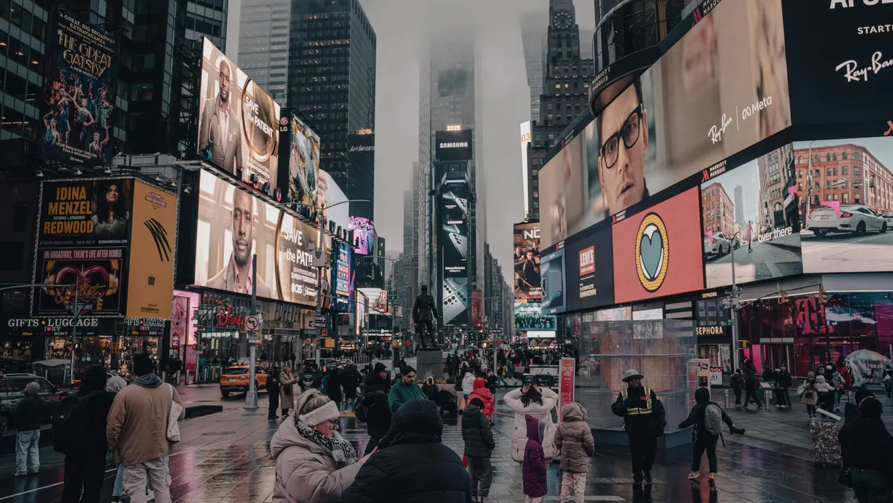 Bustling Times Square in overcast weather features vibrant billboards, a mixture of advertisements and pedestrians, creating a lively urban atmosphere.