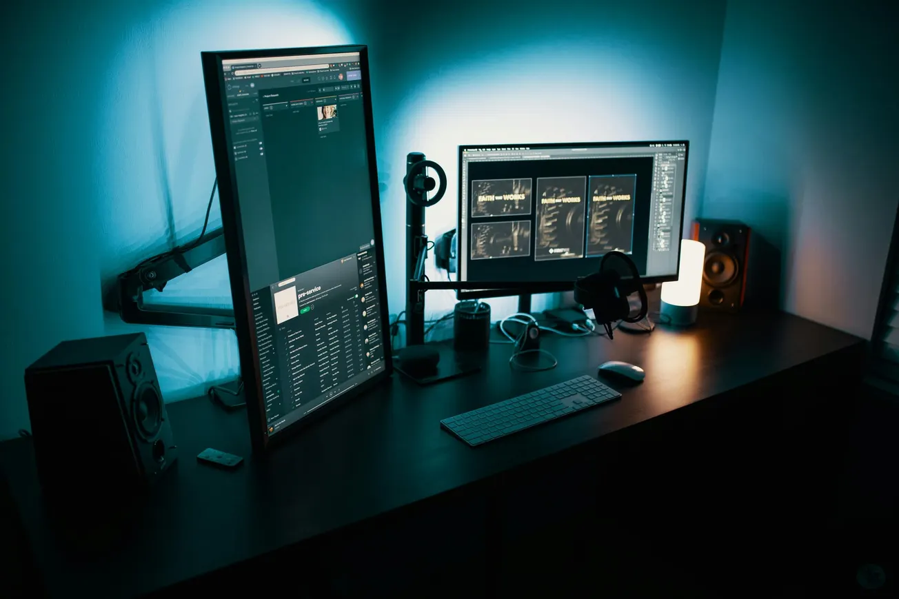 A modern home office setup features an L-shaped desk, vertically-aligned monitor, horizontal screen, keyboard, speakers, and warm desk lamp in a dim blue-lit room.