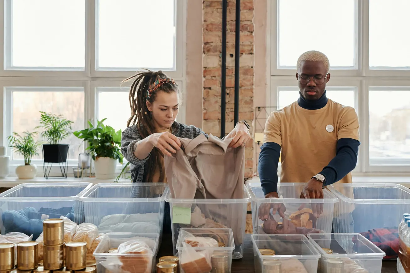 Two people sorting clothes and food items into clear bins in a bright room with large windows. The scene conveys teamwork and community service.