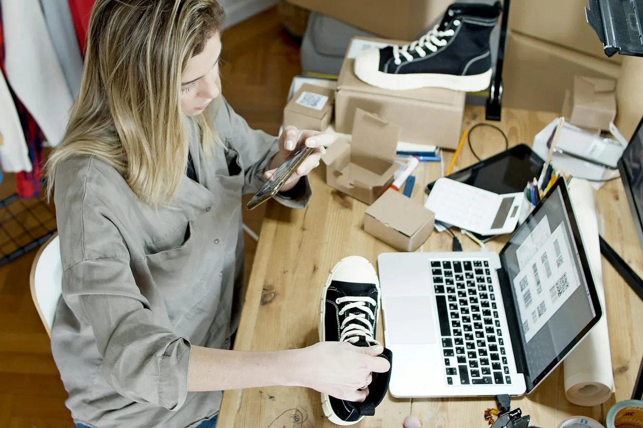 A woman in a casual gray shirt photographs black sneakers at a cluttered wooden desk with a laptop, boxes, and papers.