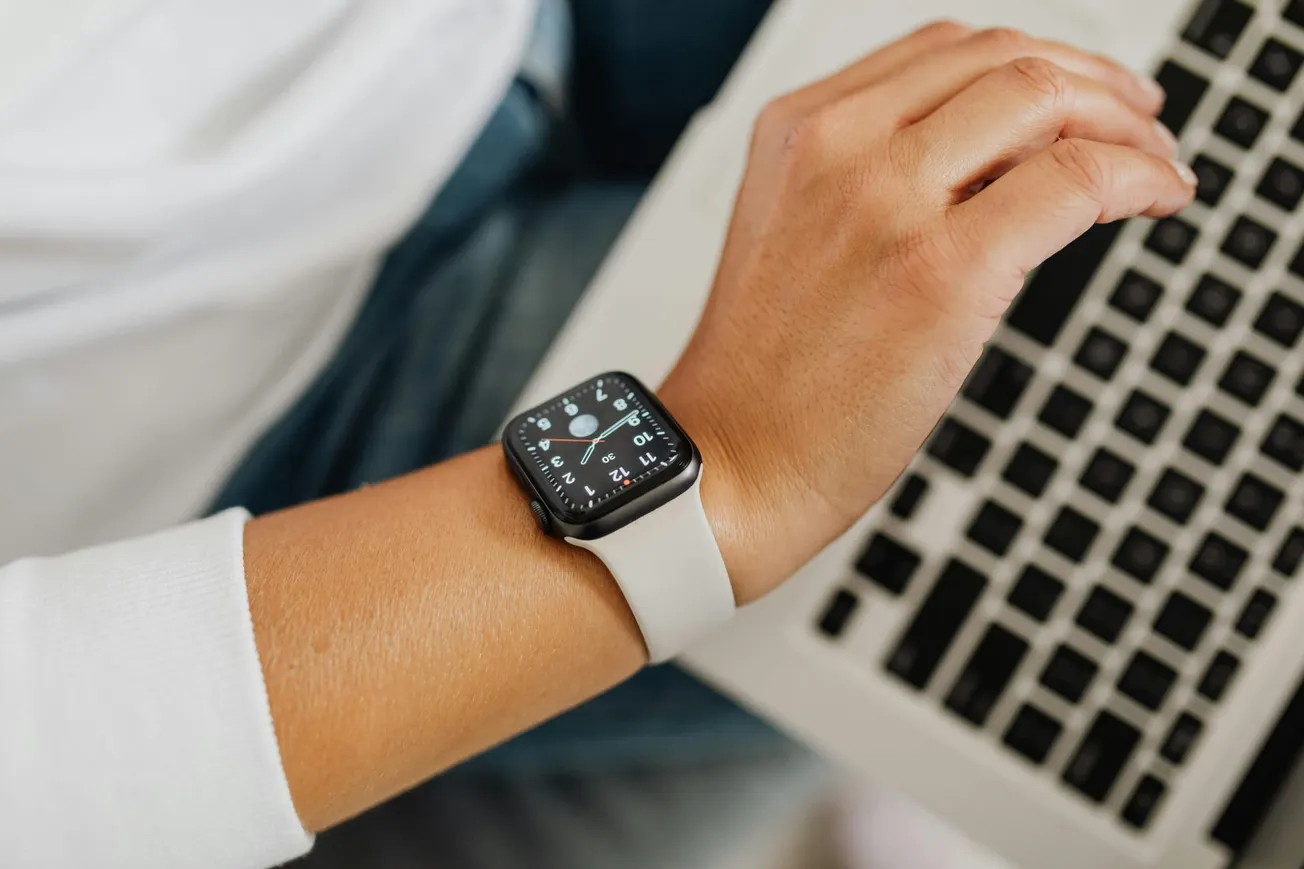 Close-up of a person's hand on a laptop keyboard, wearing a smartwatch with a white band. The watch face shows the time. The scene is casual and modern.