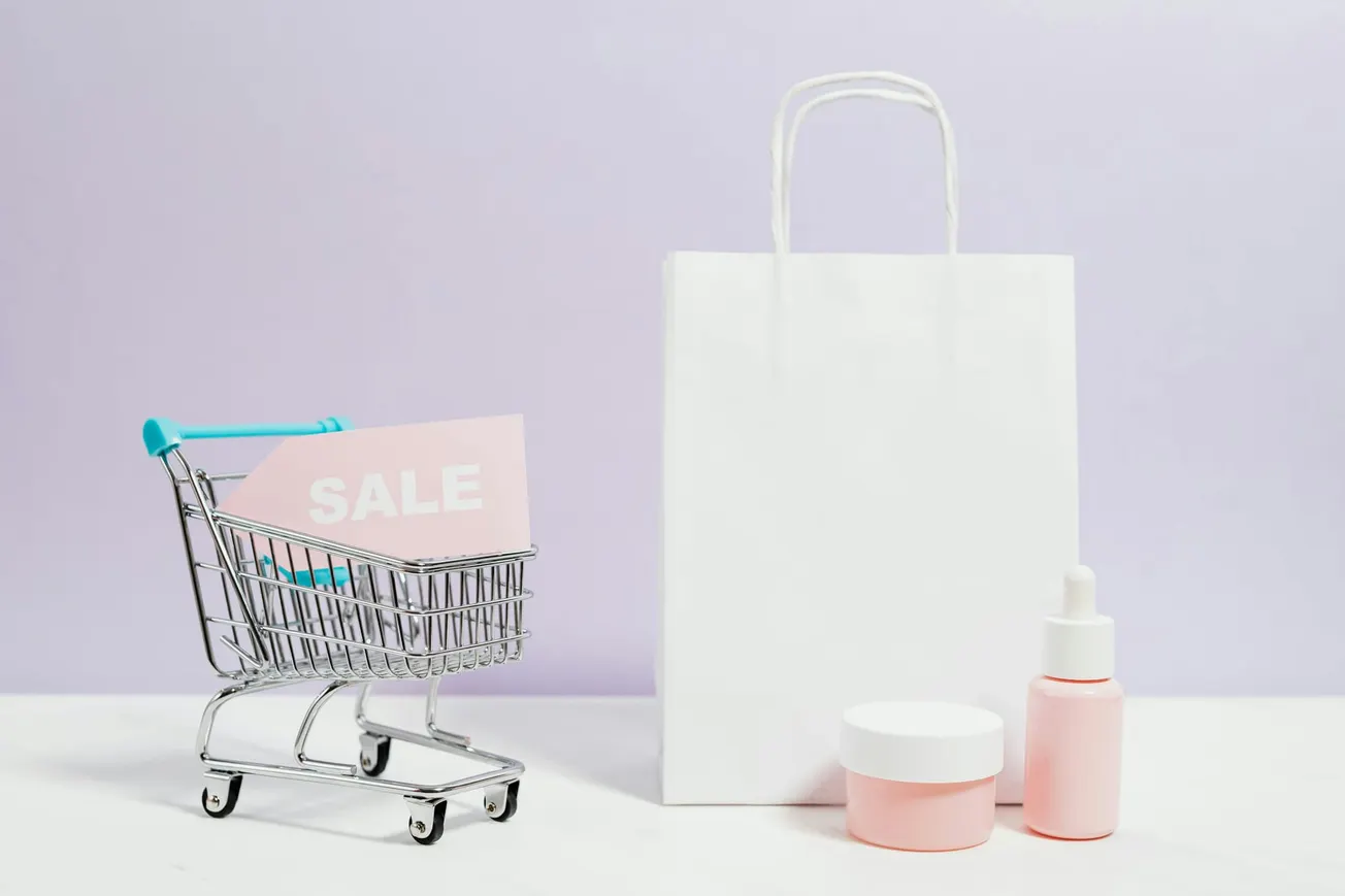 Mini shopping cart with "SALE" sign, white shopping bag, and pink cosmetic bottles on a light purple background, conveying a retail sale theme.