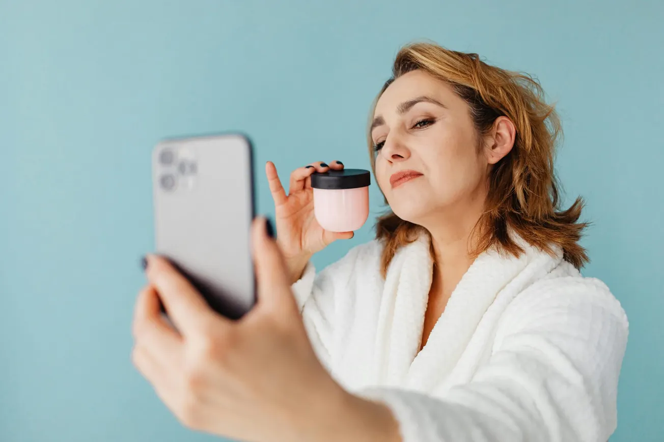 Woman in a white robe taking a selfie with a phone, holding a pink jar near her face against a blue background. She appears content and confident.